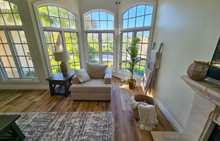 A comfortable living room with a chaise lounge, large windows offering scenic views, and decorative elements like plants and a fireplace mantel.