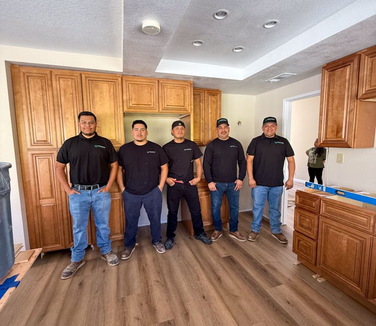 A group of renovation professionals dressed in uniform, standing together in a kitchen under construction with wooden cabinets and modern lighting.