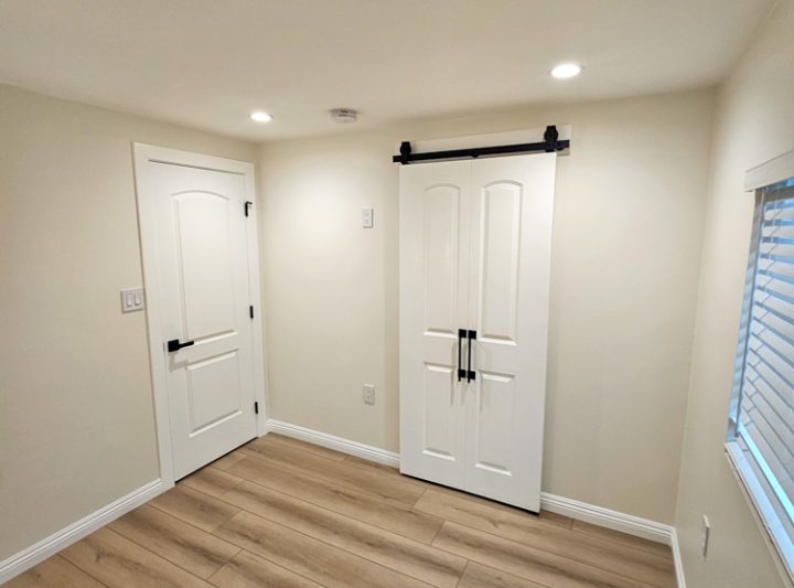 A bright room with two white doors, one with a barn-style design, beige walls, and a window with blinds allowing natural light to enter.